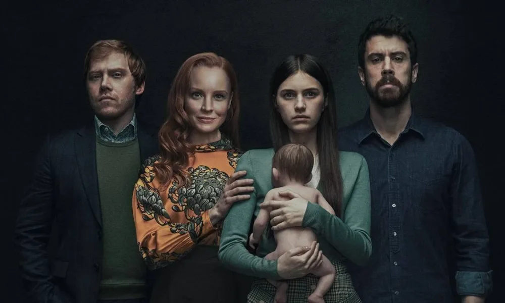 Quatro personagens da série "Servant" posando em família. A segunda mulher, ruiva, sorri, os demais membros da família estão com uma feição preta. O fundo é preto. A segunda mulher segura um bebê reborn desnudo virado com o lado do rosto para seu peito, apoiando seu colo e costas em sua mão.