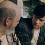 Trailer com David Corenswet, à direita, como o Superman no novo filme, homônimo, do herói, por James Gunn. Ele está sentado e como Clark Kent, conversando com um homem à esquerda, ambos de cabeça virada um ao outro.