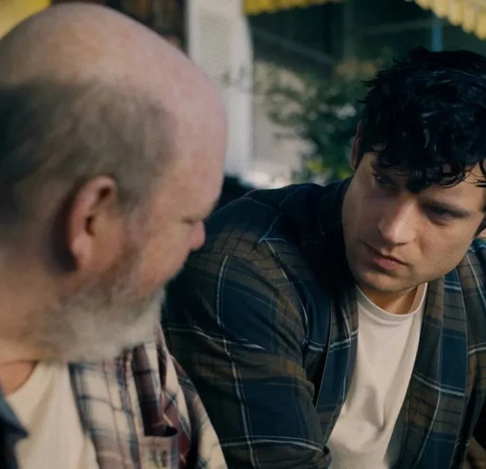 Trailer com David Corenswet, à direita, como o Superman no novo filme, homônimo, do herói, por James Gunn. Ele está sentado e como Clark Kent, conversando com um homem à esquerda, ambos de cabeça virada um ao outro.