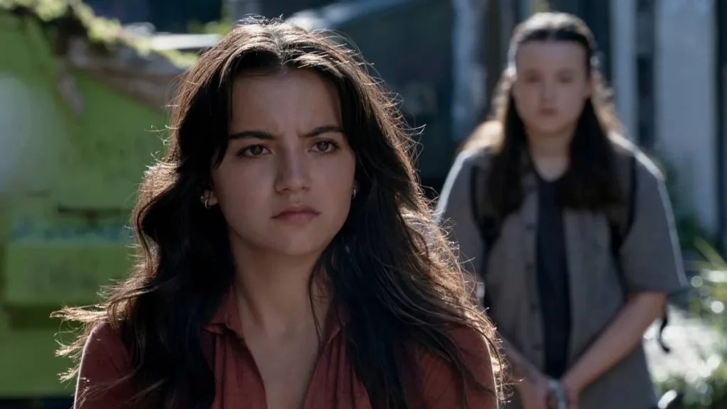 The Last of Us | 2ª Temporada Ameaça Ficar Boa e É Inocentada Por Falta de Provas 2 Dina (esquerda), interpretada por Isabela Merced, em destaque, com um semblante sério e levemente irritado em 'The last of Us". Ellie (Bella Ramsey) está atrás dela, em guarda com uma arma, desfocada.