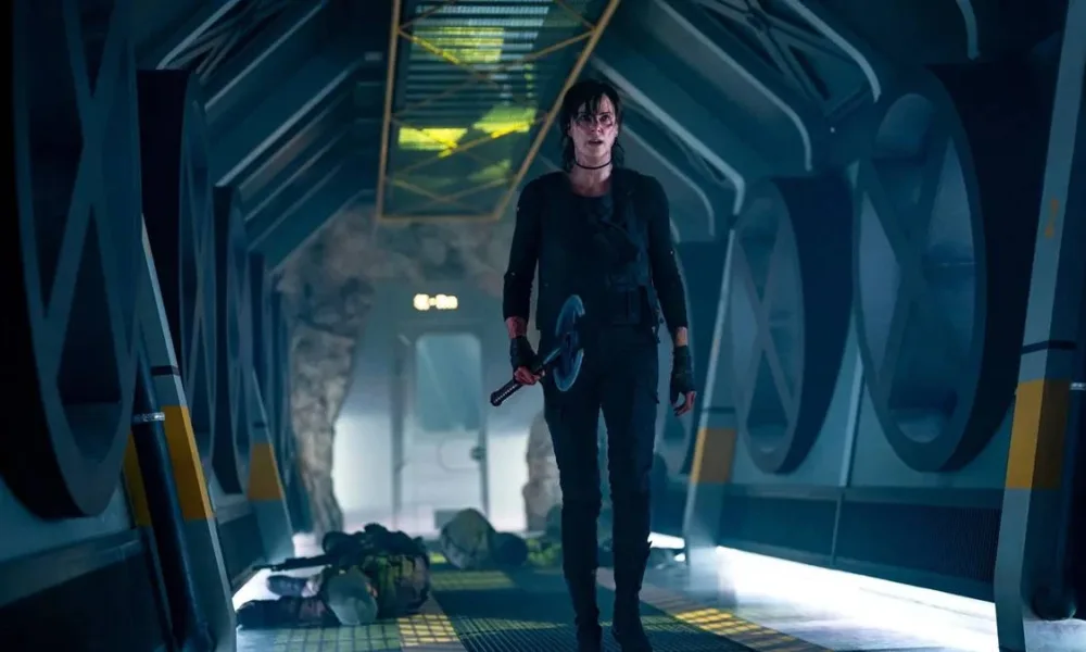 Charlize Theron como Andy em "The Old Guard 2", em uma nave, empunhando um machado com a mão direita, e vários corpos atrás dela, recém caídos.