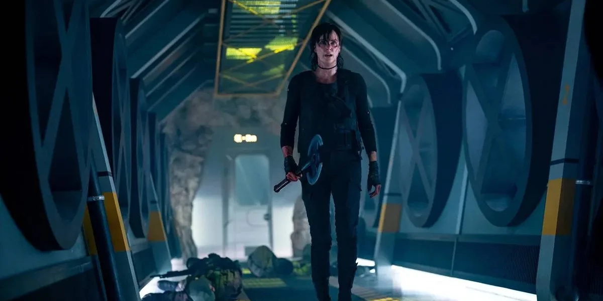 Charlize Theron como Andy em "The Old Guard 2", em uma nave, empunhando um machado com a mão direita, e vários corpos atrás dela, recém caídos.
