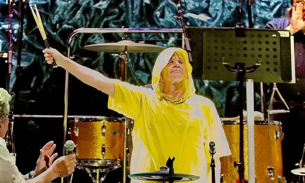 Baterista Zak Starkey durante show do The Woo, vestindo uma capa de chuva amarela e com braço direito (nossa esquerda) erguido em 45º segurando as baquetas.