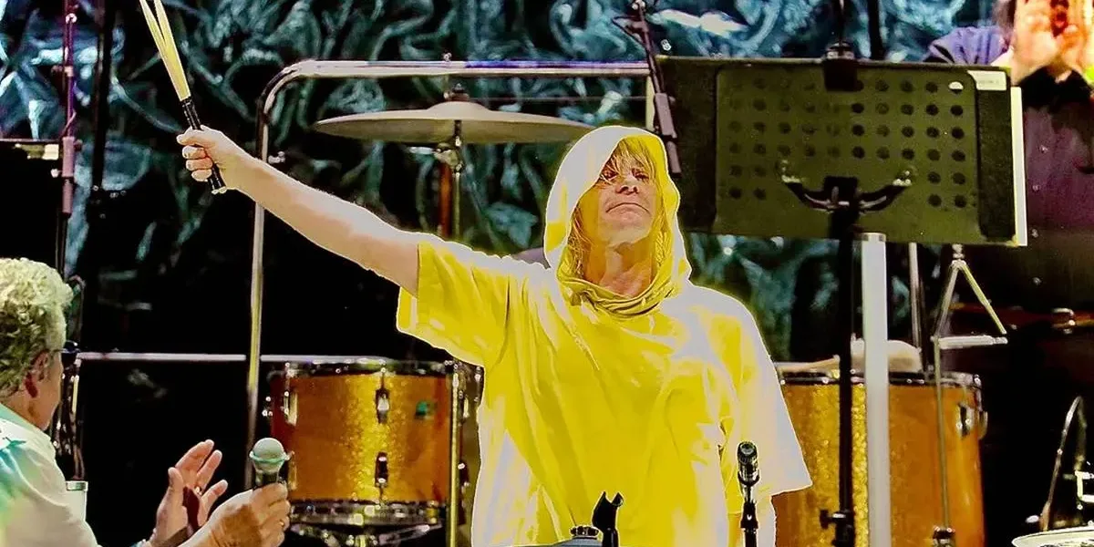 Baterista Zak Starkey durante show do The Woo, vestindo uma capa de chuva amarela e com braço direito (nossa esquerda) erguido em 45º segurando as baquetas.