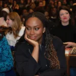 Autora Chimamanda Adichie, sorrindo e olhando de canto, vestida de preto e sentada em uma plateia na Sciences Po University. Há várias mulheres em destaque na foto.
