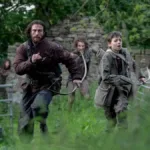 Jamie (Aaron Taylor-Johnson) e Spike (Alfie Williams) fugindo de três zumbis ao fundo deles em "Extermínio: A Evolução". Ambos carregam arcos e flechas.