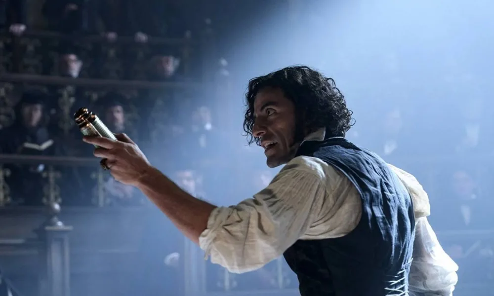 Oscar Isaac com cabelo longo e solto, ondulado, interpretando o Dr. Victor Frankenstein na versão de Guillermo del Toro do clássico "Frankenstein". Ele está com braço direito (a nossa esquerda) levantado em arco e segurando, apontando-o, um frasco dourado.