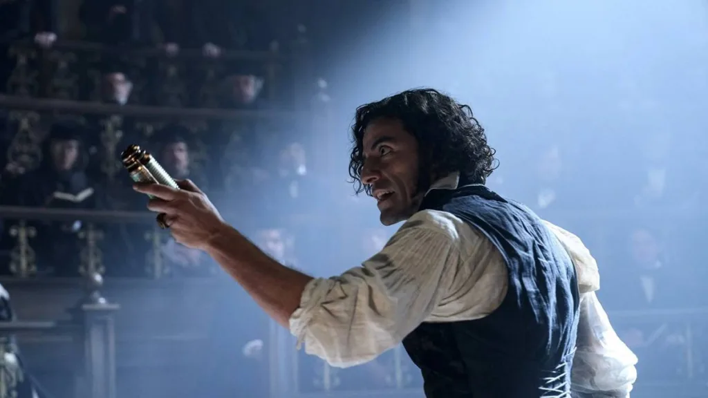 Oscar Isaac com cabelo longo e solto, ondulado, interpretando o Dr. Victor Frankenstein na versão de Guillermo del Toro do clássico "Frankenstein". Ele está com braço direito (a nossa esquerda) levantado em arco e segurando, apontando-o, um frasco dourado.