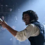 Oscar Isaac com cabelo longo e solto, ondulado, interpretando o Dr. Victor Frankenstein na versão de Guillermo del Toro do clássico "Frankenstein". Ele está com braço direito (a nossa esquerda) levantado em arco e segurando, apontando-o, um frasco dourado.