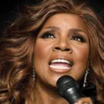 Gloria Gaynor em 2024 para o documentário musical "I Will Survive". Ela canta olhando para o canto superior esquerdo da tela, focalizado em seu rosto, em um fundo preto.