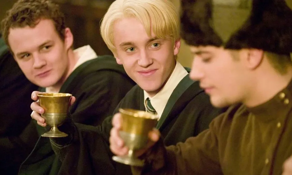 Tom Felton em destaque como Draco Malfoy, trajado com as roupas de Hogwarts, observando o aluno Viktor Krum, todos bebendo de seus cálices no quarto filme de "Harry Potter", o "cálice de fogo".