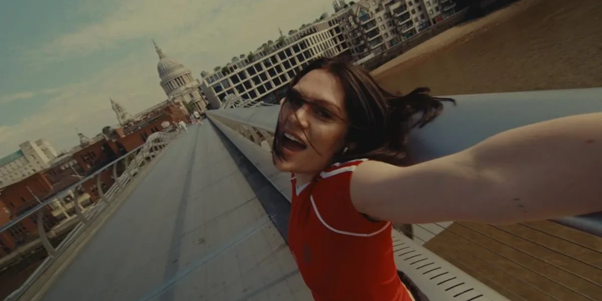 Câmera panorâmica filmando Jessie J, de braços abertos e sorrindo em uma ponte, de camiseta vermelha com duas listas na altura do ombro no clipe de "Living My Best Life" (2025).