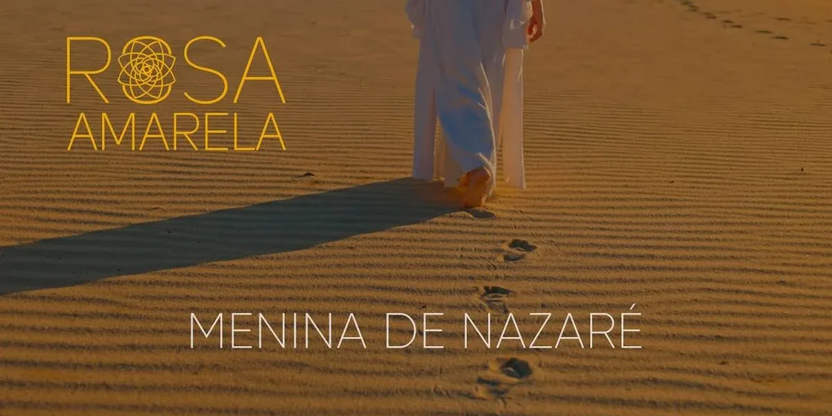 Rosa Amarela Lança “Menina de Nazaré” em Videoclipe, Evocando Fé e Reconexão Capa do clipe "Menina de Nazaré", do duo Rosa Amarela. Pris Mariano, no lado direito do quadrante, quase ao meio, caminha nas areias e deixa pegadas. Está de dia e podemos ver sua sombra do lado esquerdo.