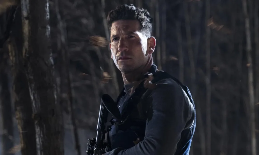 A imagem mostra Jon Bernthal interpretando Frank Castle, também conhecido como O Justiceiro (The Punisher). Ele está em um bosque escuro de noite, olhos semicerrados, segurando uma grande arma no peito e de colete.