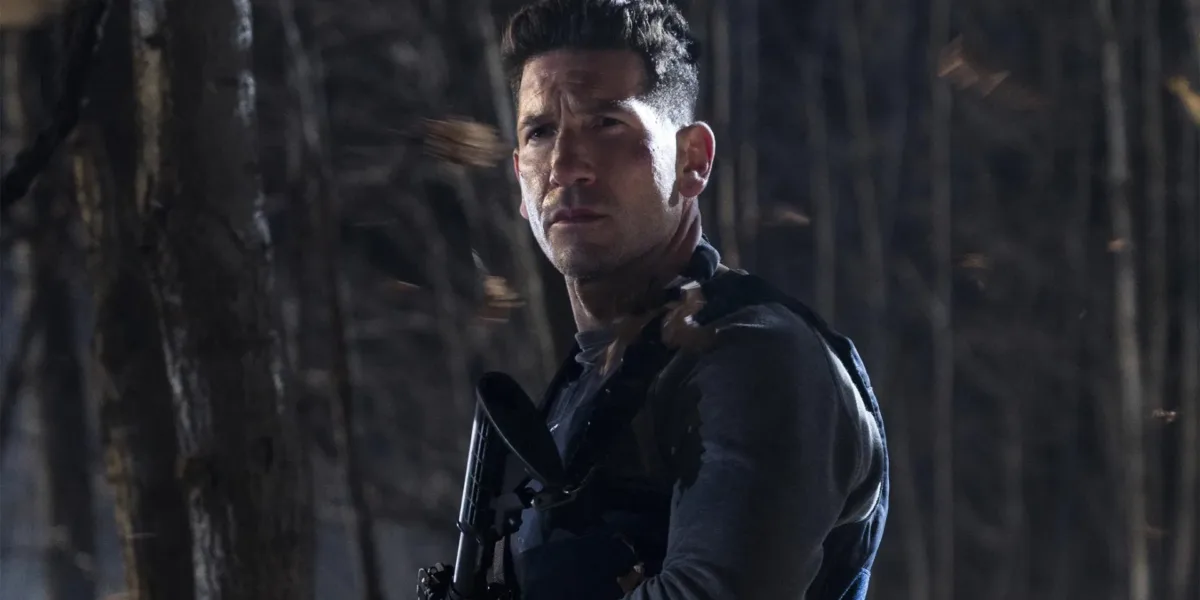A imagem mostra Jon Bernthal interpretando Frank Castle, também conhecido como O Justiceiro (The Punisher). Ele está em um bosque escuro de noite, olhos semicerrados, segurando uma grande arma no peito e de colete.