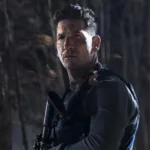 A imagem mostra Jon Bernthal interpretando Frank Castle, também conhecido como O Justiceiro (The Punisher). Ele está em um bosque escuro de noite, olhos semicerrados, segurando uma grande arma no peito e de colete.