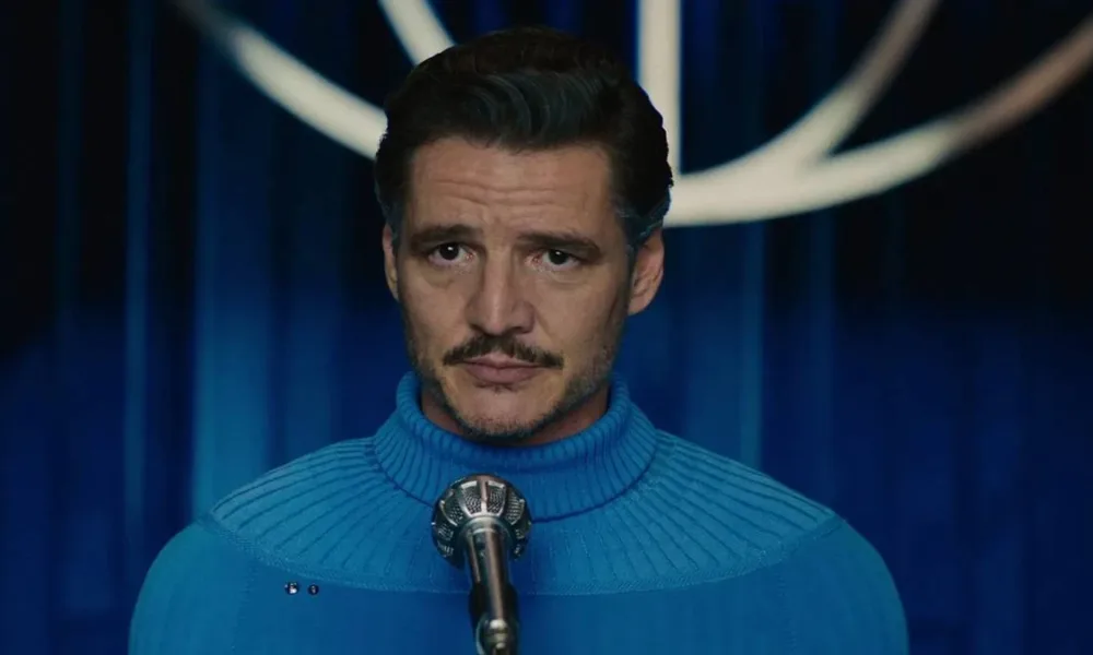 Pedro Pascal com suéter de gola alta azul da cor do uniforme do Quarteto Fantástico, no filme de mesmo nome, 2025, onde interpreta o Senhor Fantástico. Ele está centralizado na tela, com um microfone em sua frente.
