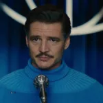 Pedro Pascal com suéter de gola alta azul da cor do uniforme do Quarteto Fantástico, no filme de mesmo nome, 2025, onde interpreta o Senhor Fantástico. Ele está centralizado na tela, com um microfone em sua frente.