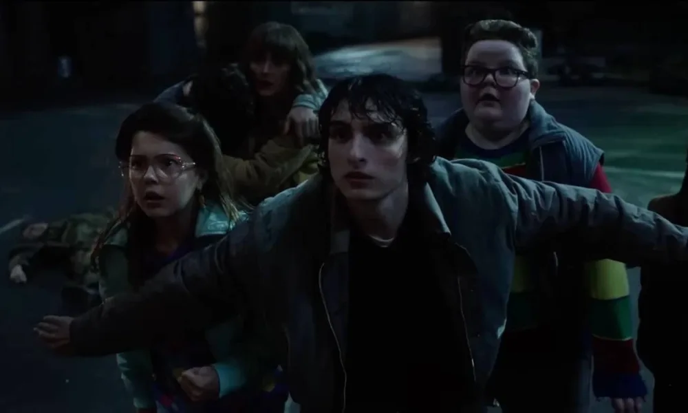 Mike, de "Stranger Things", com os braços abertos para trás segurando um grupo de cidadãos, majoritariamente crianças, no sétimo episódio da 5ª temporada da série. Todos estão assustados, e a mãe de Will (Winona Ryder) está presente também.