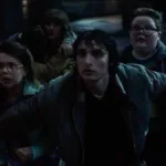 Mike, de "Stranger Things", com os braços abertos para trás segurando um grupo de cidadãos, majoritariamente crianças, no sétimo episódio da 5ª temporada da série. Todos estão assustados, e a mãe de Will (Winona Ryder) está presente também.