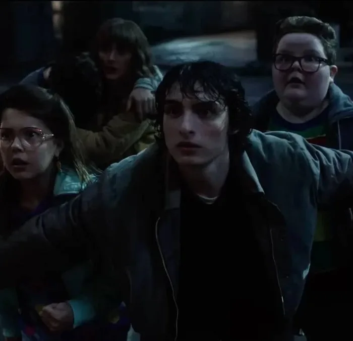 Mike, de "Stranger Things", com os braços abertos para trás segurando um grupo de cidadãos, majoritariamente crianças, no sétimo episódio da 5ª temporada da série. Todos estão assustados, e a mãe de Will (Winona Ryder) está presente também.