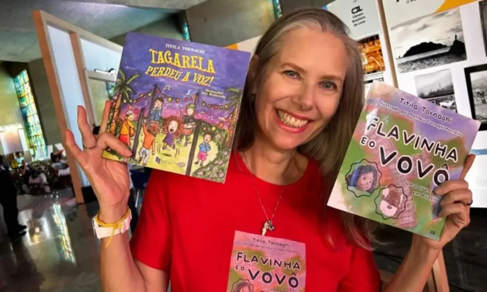 Autora Titila Tornaghi na Bienal do Livro Rio 2025 segurando dois livros seus e sorrindo: à nossa esquerda "Tagarela perdeu a voz!" e à nossa direita "Flavinha e o vovô", do qual veste uma camisa vermelha que estampa a capa também.