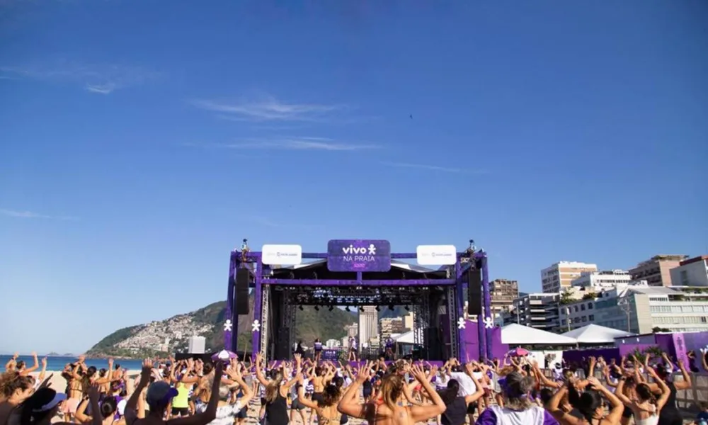Campanha Vivo na Praia, no Rio de Janeiro, com um grande palco roxo centralizado em um dia de sol e a praia lotada.