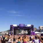 Campanha Vivo na Praia, no Rio de Janeiro, com um grande palco roxo centralizado em um dia de sol e a praia lotada.