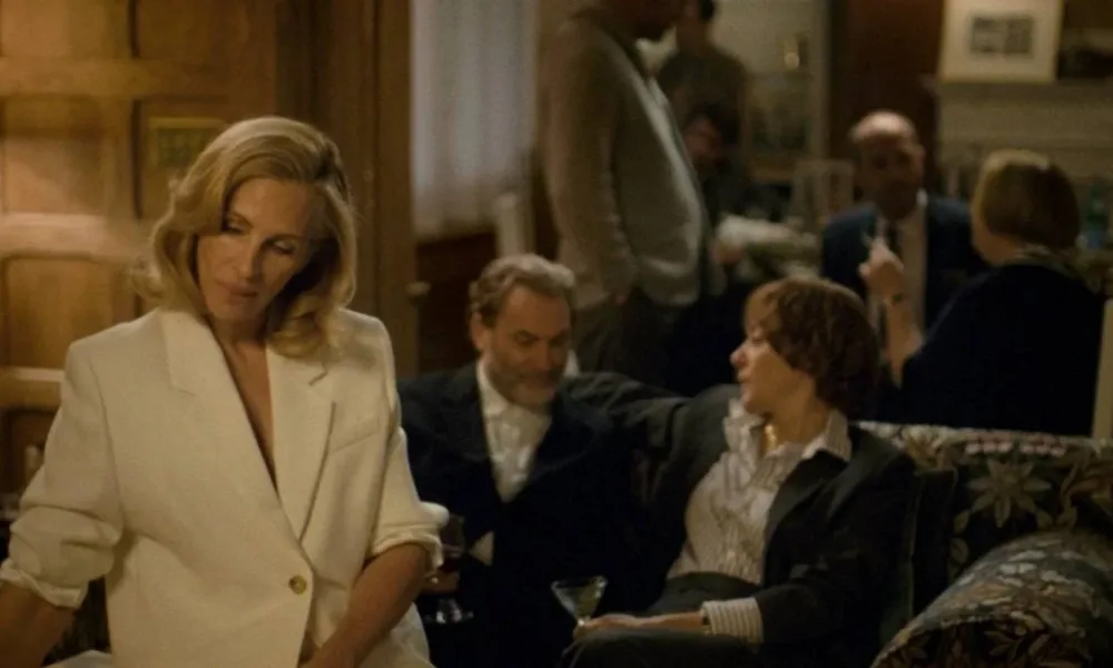 De branco, sentada no braço de um sofá, Julia Roberts é Alma Olsson, protagonista de "Depois da Caçada", em cena com encontro da alta sociedade no filme.