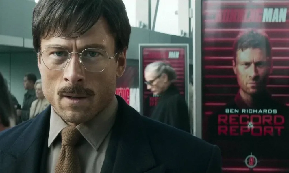 Glen Powell em "O Concorrente", remake de 2025. Ele está de terno e gravata, óculos e bigode, e uma expressão de angústia.
