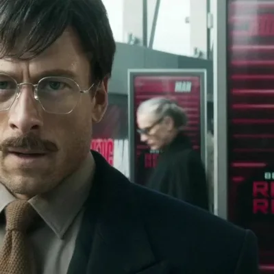 Glen Powell em "O Concorrente", remake de 2025. Ele está de terno e gravata, óculos e bigode, e uma expressão de angústia.