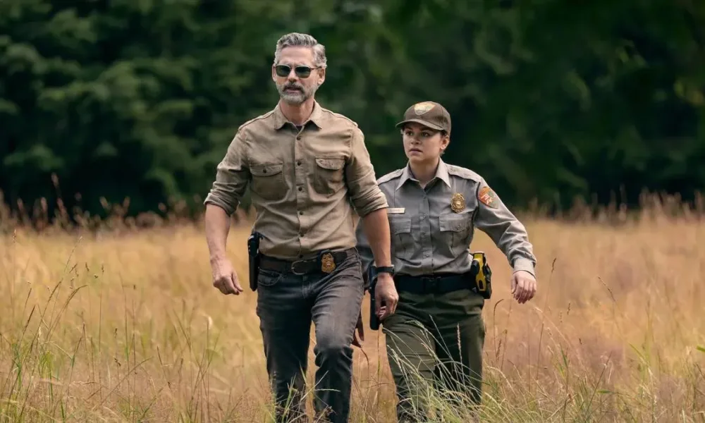 Eric Bana e Lily Santiago como Kyle Turner e Naya Vazques em meio a grama alta seca e vestidos como agentes na série da Netflix "Indomável".