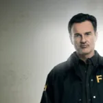 Julian McMahon como Jess LaCroix, seu personagem em "FBI: Os Mais Procurados", fardado e frente a um fundo cinza com luz irradiando atrás dele.