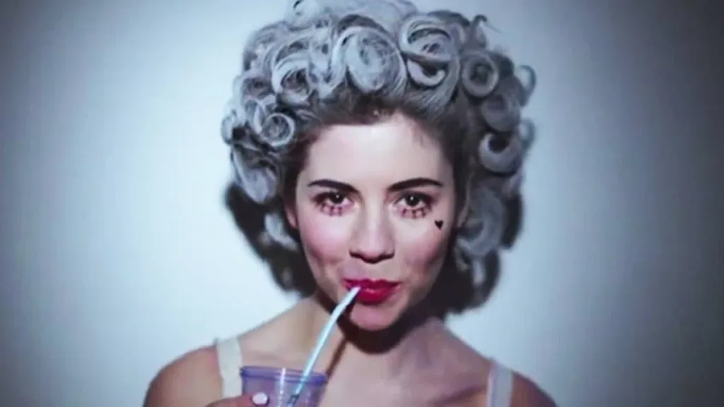 Princess of Power — Marina | Sobre Se Tornar Caricatura de Si Mesmo 1 Thumbnail de "Primadonna", do segundo álbum de estúdio de Marina and the Diamonds. Ela está com uma peruca cinza com penteado dos anos 50, maquiagem pálida, um coração desenhado na bochecha, sorrindo maliciosamente enquanto bebe uma bebida de canudo.