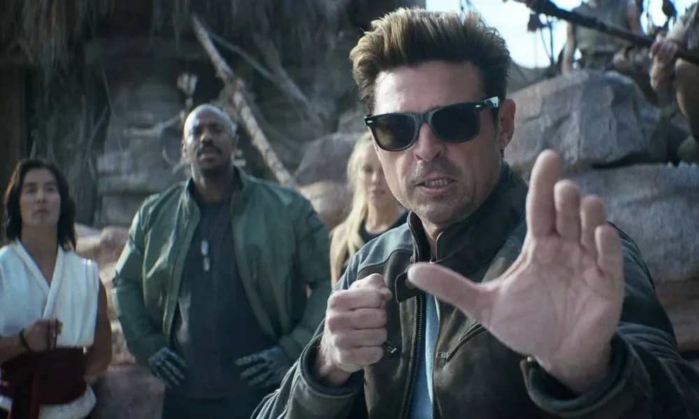 Karl Urban à direita em destaque, de óculos, como Johnny Cage no trailer de "Mortal Kombat 2", em posição de combate com a mão esquerda a frente do corpo.