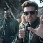 Karl Urban à direita em destaque, de óculos, como Johnny Cage no trailer de "Mortal Kombat 2", em posição de combate com a mão esquerda a frente do corpo.