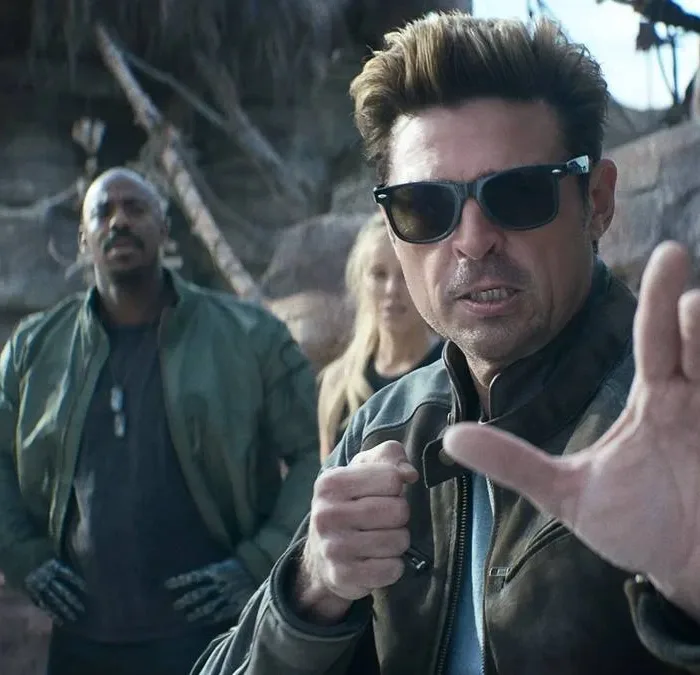 Karl Urban à direita em destaque, de óculos, como Johnny Cage no trailer de "Mortal Kombat 2", em posição de combate com a mão esquerda a frente do corpo.