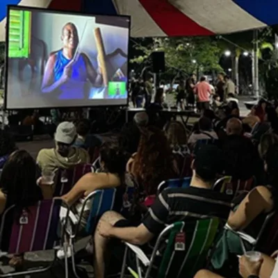 Exibição de filme pelo projeto Faz na Praça. Pessoas assistem a película em cadeiras de sol sob uma lona, em espaço aberto.