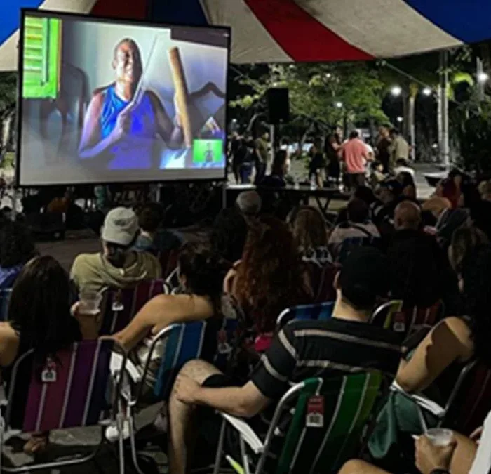 Exibição de filme pelo projeto Faz na Praça. Pessoas assistem a película em cadeiras de sol sob uma lona, em espaço aberto.