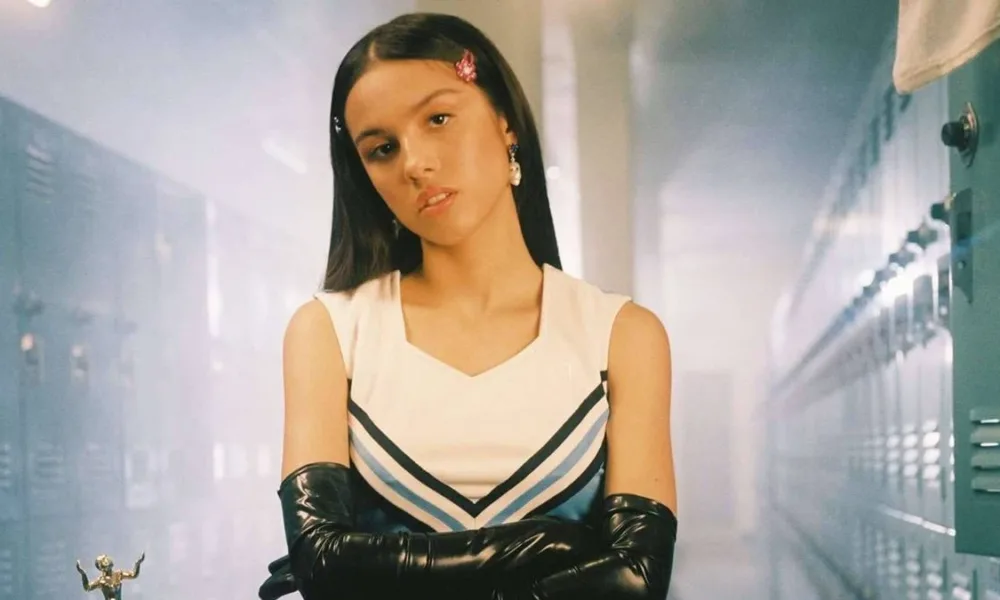 Olivia Rodrigo, cara blasé, segurando os braços com suas luvas compridas de látex pretas, e uniforme de colegial em um corredor de highschool no clipe de "Good 4u".