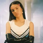 Olivia Rodrigo, cara blasé, segurando os braços com suas luvas compridas de látex pretas, e uniforme de colegial em um corredor de highschool no clipe de "Good 4u".