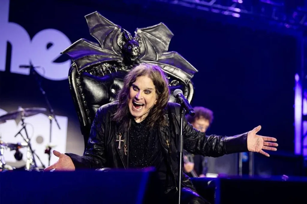 Back to the Beginning | Confira as Músicas que Estarão na Versão Cinematográfica da Despedida de Ozzy Osbourne 1 Foto de Ozzy em trono negro adornado com um morcego, o vocalista de braços abertos sorrindo de boca aberta provocativamente, maquiagem forte, cabelos longos, em foto promocional para o "Back To The Beginning: Ozzy’s Final Bow", que será lançado em 2026.