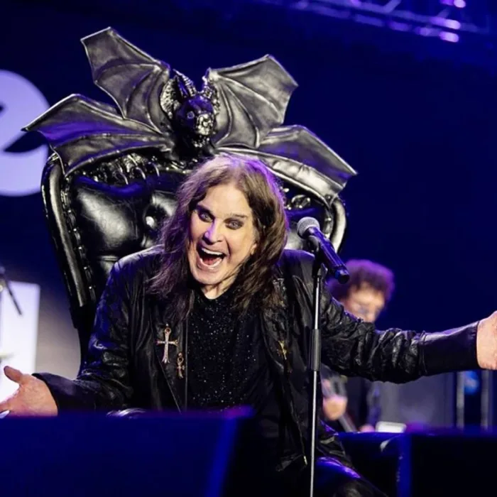 Foto de Ozzy em trono negro adornado com um morcego, o vocalista de braços abertos sorrindo de boca aberta provocativamente, maquiagem forte, cabelos longos, em foto promocional para o "Back To The Beginning: Ozzy’s Final Bow", que será lançado em 2026.