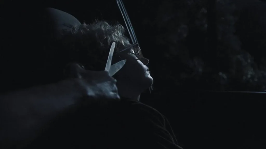 A Hora do Mal | O Maior Terror É O Desconhecido 2 Julia Garner como Justine em "A Hora do Mal", deitada em um carro no escuro, de noite. Uma mão masculina se aproxima dela com uma tesoura.
