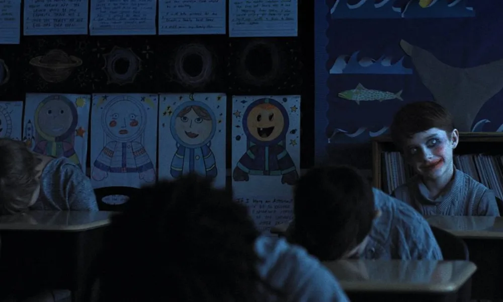 Crianças em sala de aula em "A Hora do Mal", de cabeça para baixo. Um garoto, ao fundo, é o único de cabeça erguida, com um sorriso desconcertante e maquiagem à Coringa.