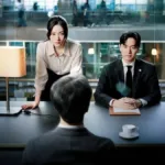 Protagonistas de "Além do Direito", os atores Jung Chae-yeon e Lee Jin-uk vestidos como advogados. Ela com as mãos apoiadas na mesa e corpo reclinado, ele com as mãos juntas e também com expressão séria. Do outro lado da mesa há um personagem masculino de costas, olhando para eles, contra a nossa câmera.