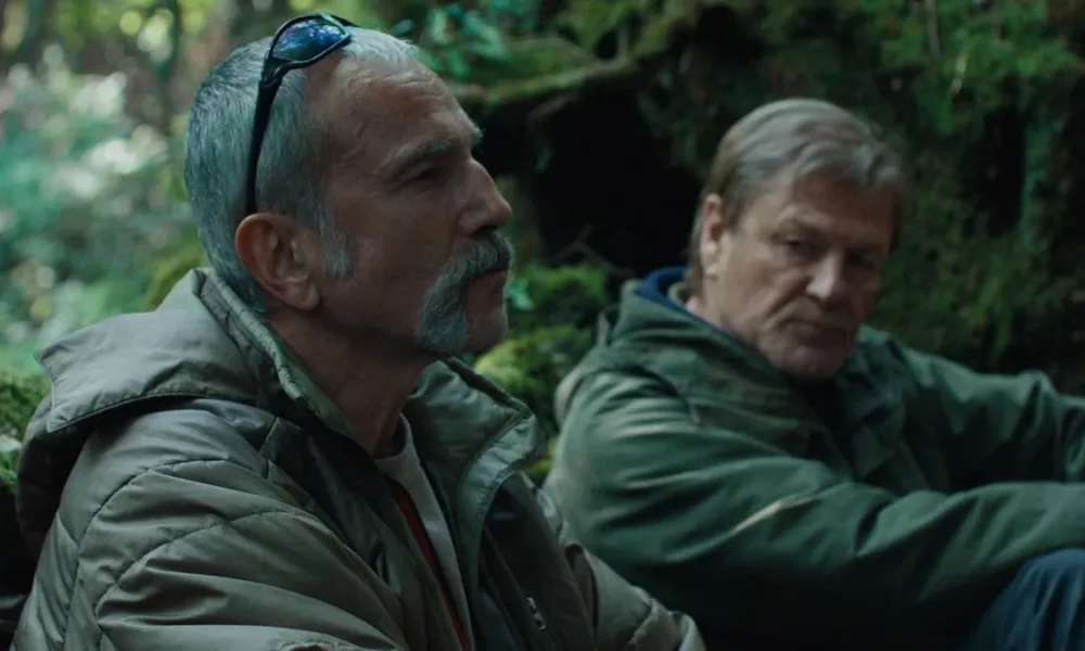 Daniel Day-Lewis e Sean Bean em uma mata aparentemente tropical no filme "Anemone", trajados de um verde militar. O protagonista está de perfil, enquanto Sean olha para baixo em direção ao protagonista.