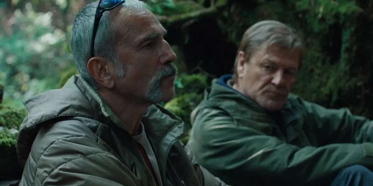 Daniel Day-Lewis e Sean Bean em uma mata aparentemente tropical no filme "Anemone", trajados de um verde militar. O protagonista está de perfil, enquanto Sean olha para baixo em direção ao protagonista.