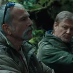 Daniel Day-Lewis e Sean Bean em uma mata aparentemente tropical no filme "Anemone", trajados de um verde militar. O protagonista está de perfil, enquanto Sean olha para baixo em direção ao protagonista.