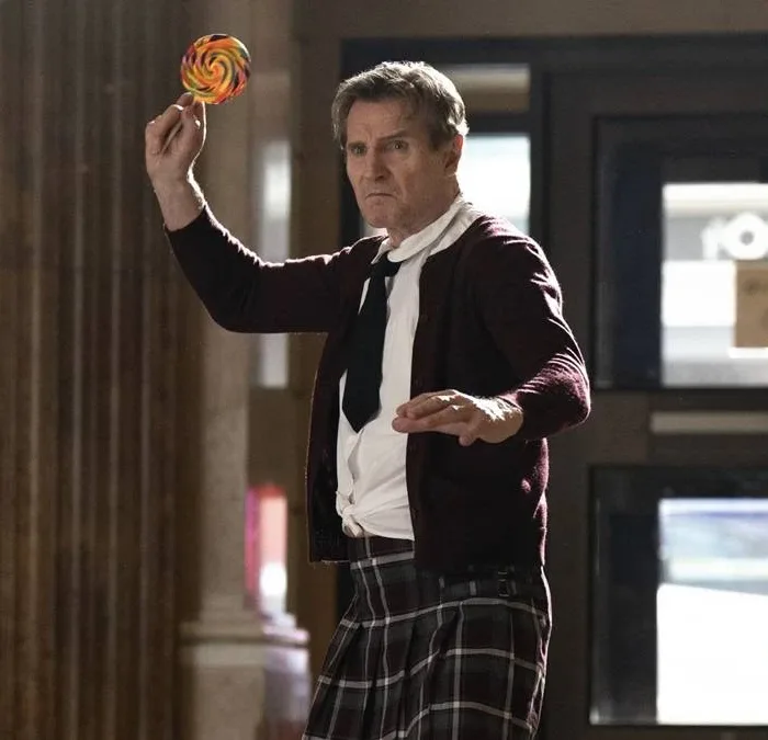 Liam Neeson vestindo kilt, roupa social e gravata, empunhando um pirulito gigante colorido como se fosse uma arma de arremesso no reboot de 2025 de "Corra Que a Polícia Vem Aí".